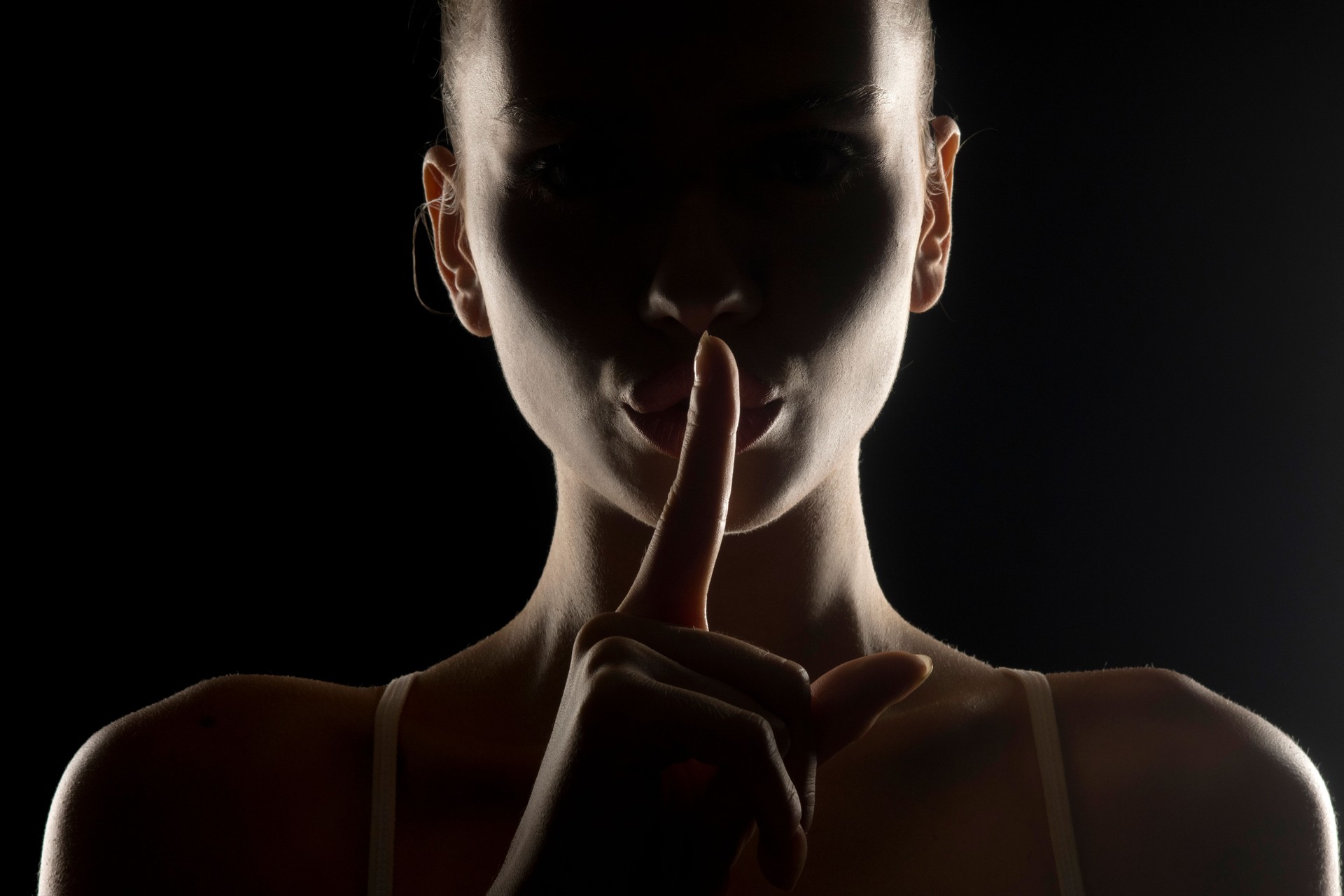 Mysterious silhouette of a woman making 'shh' gesture with finger on lips. Dark background with rim lighting. Enigmatic figure symbolizing silence or secrecy