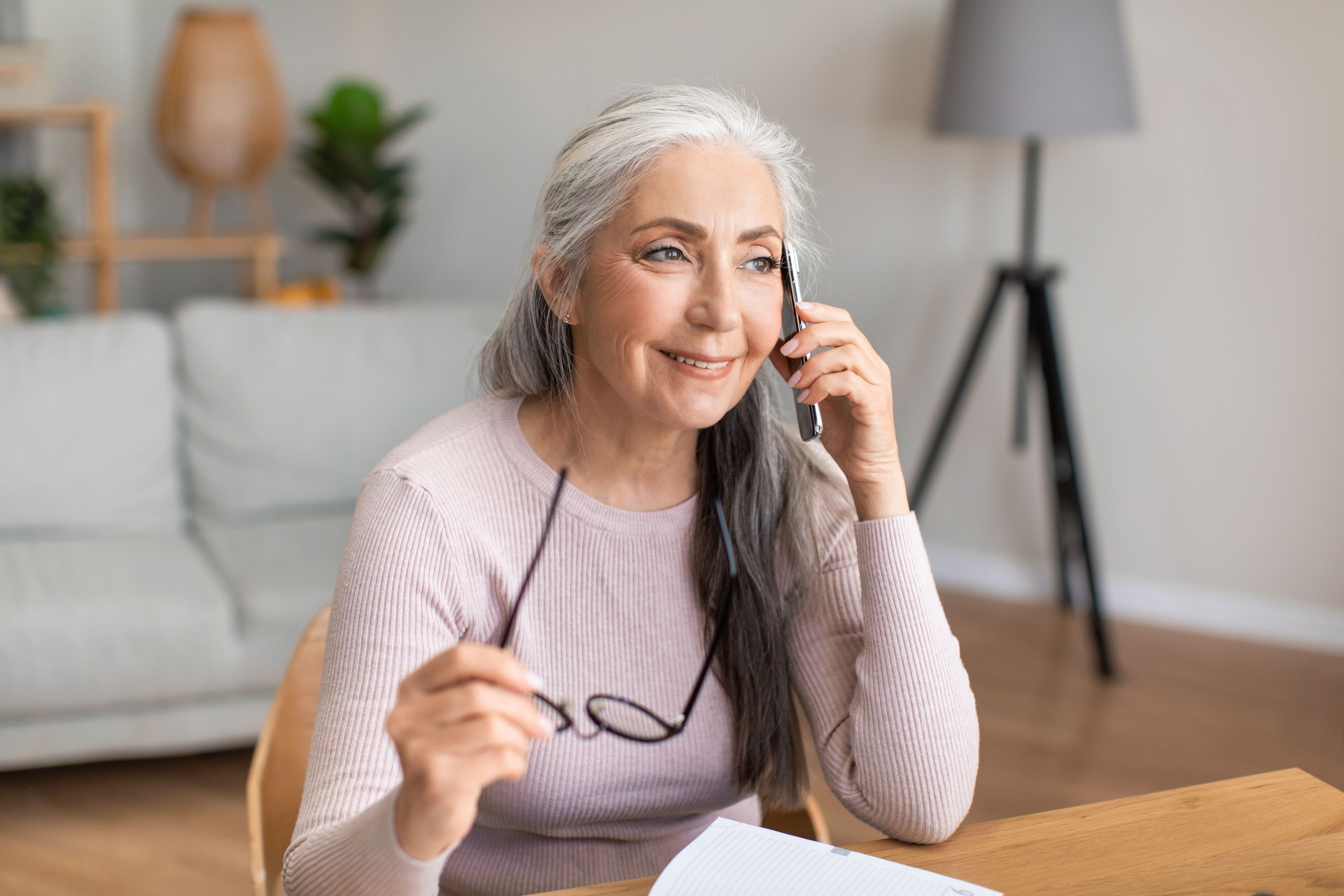 Smiling caucasian mature gray-haired lady hold glasses speaks by phone with client in living room interior
